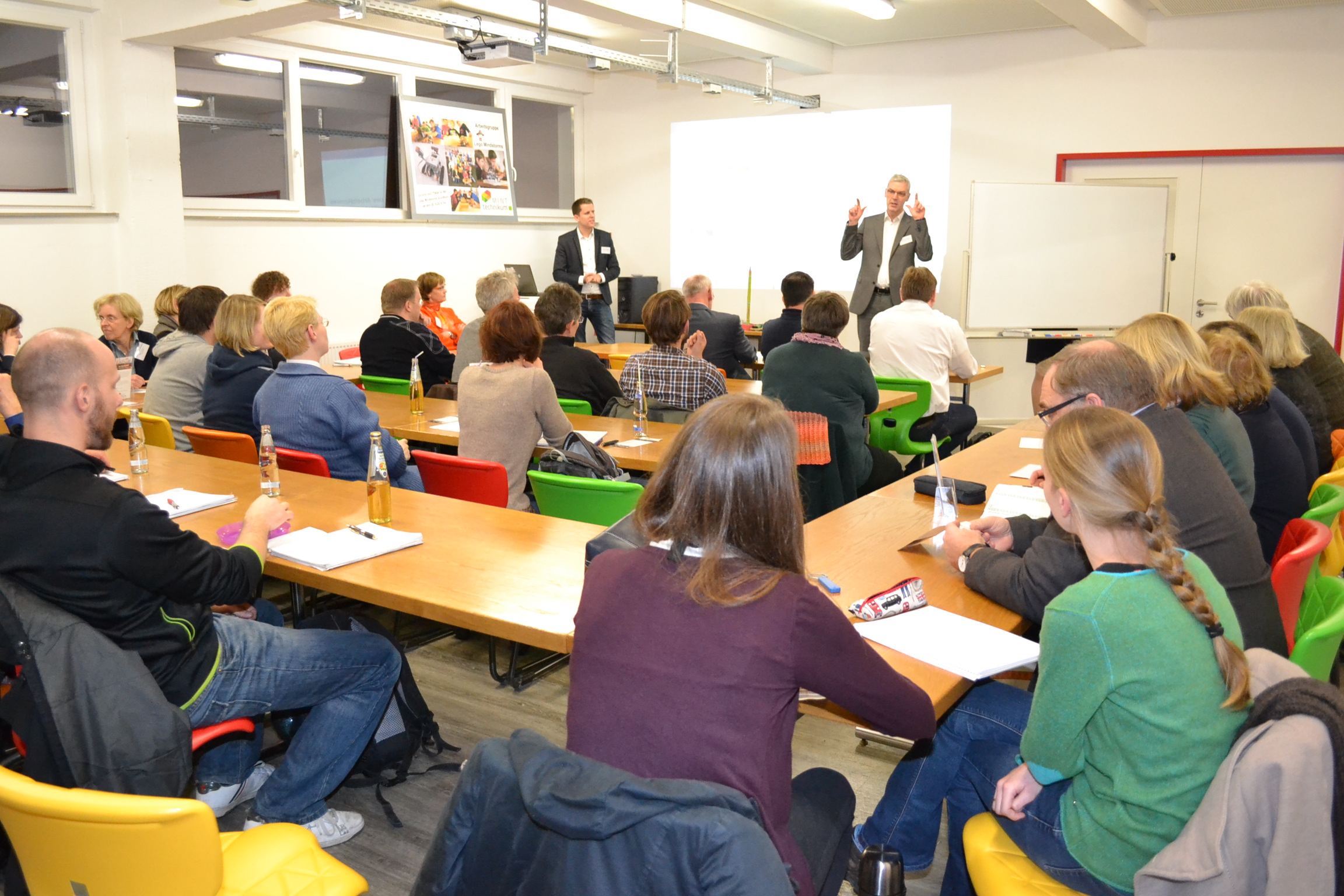 Eine Gruppe Erwachsener sitzt an Tischen in einem Klassenzimmer. Ein Mann spricht vorne, während ein anderer daneben steht. Sie diskutieren über Initiativen zur Mädchenförderung.