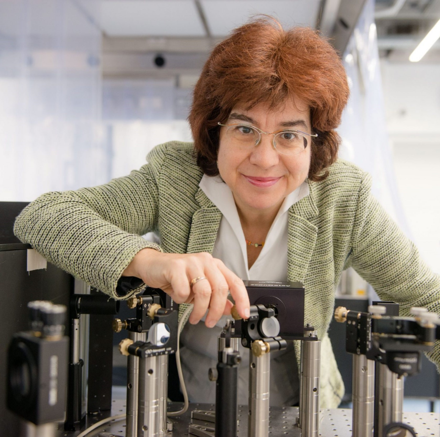 Eine Frau mit Brille und braunen Haaren stellt in einem Labor Geräte ein. Sie ist von wissenschaftlichen Instrumenten und Metallteilen umgeben.