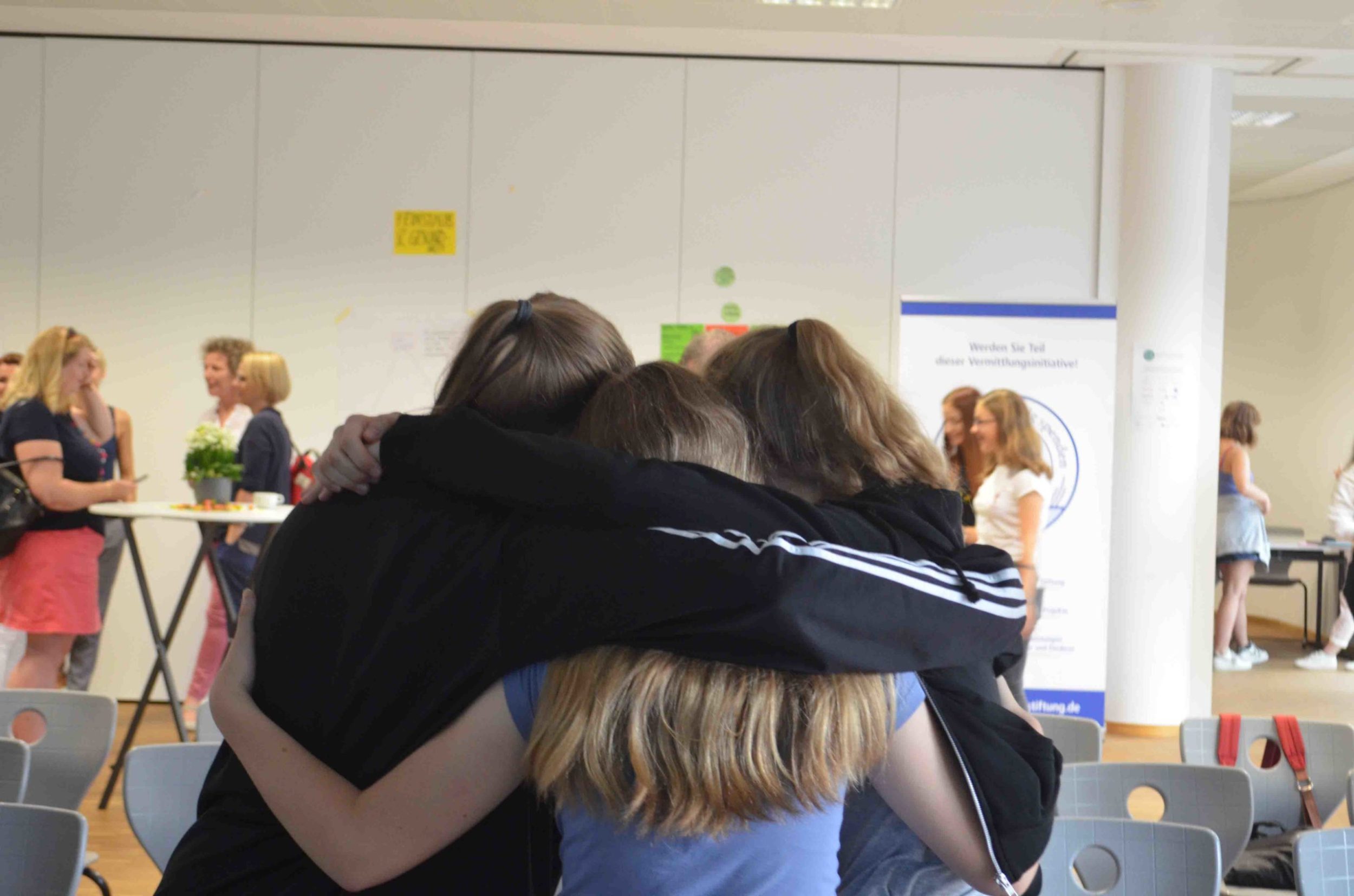 Drei Personen stehen mit umeinander gelegtem Arm in einer ungezwungenen Umgebung in einem Innenraum und blicken von der Kamera weg. Im Hintergrund unterhalten sich andere Personen, möglicherweise bei einer Veranstaltung von „girls for innovation Köln“.