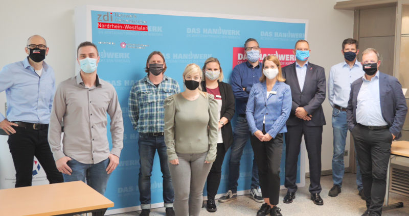 Elf Erwachsene stehen in der zdi-Handwerkstatt, tragen Mund-Nasen-Schutz und posieren vor einem blauen „Das Handwerk“-Hintergrund für ein Gruppenfoto.