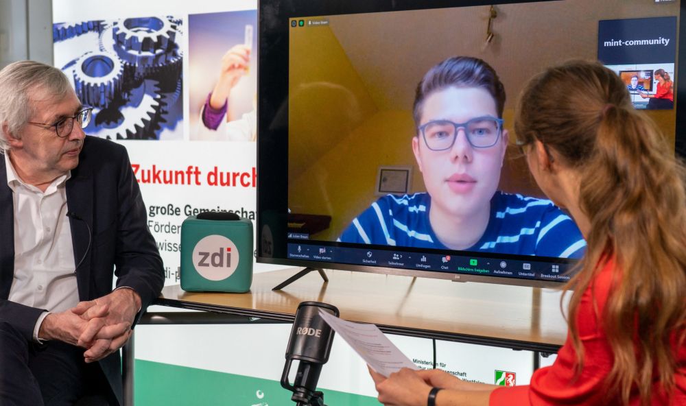 Drei Personen nehmen an einer Hybrid-Sitzung der Start MINT-Community NRW teil. Ein Mann und eine Frau sind persönlich anwesend, ein weiterer Mann ist per Videokonferenz auf einem großen Bildschirm zu sehen.