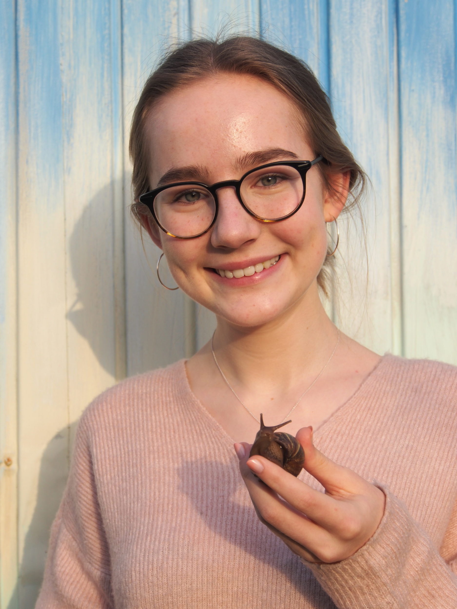 Ein junger Mensch mit Brille lächelt in die Kamera und hält eine kleine braune Schnecke in der Hand. Im Hintergrund sind hellblaue Holzbretter zu sehen. Auf dem Bild ist Marike Skiba zu sehen.
