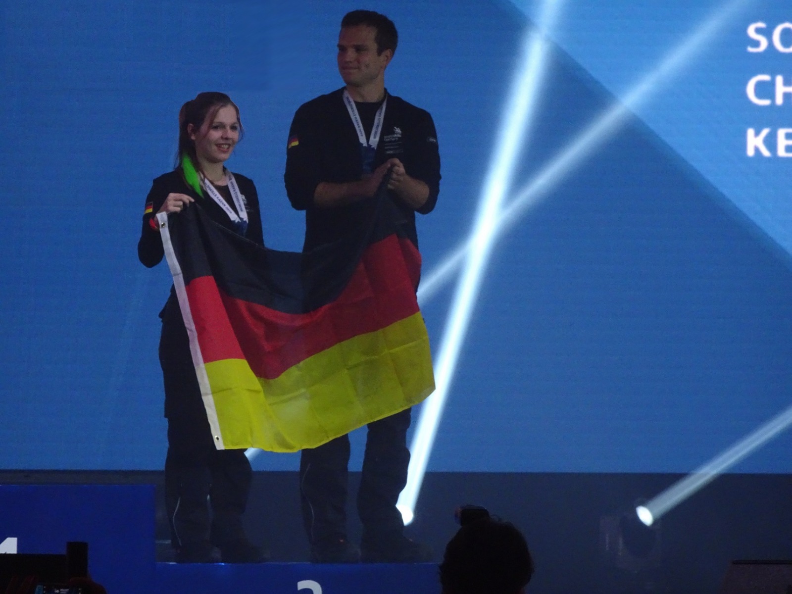 Eine Frau und ein Mann halten auf einer Bühne eine Deutschlandfahne hoch.