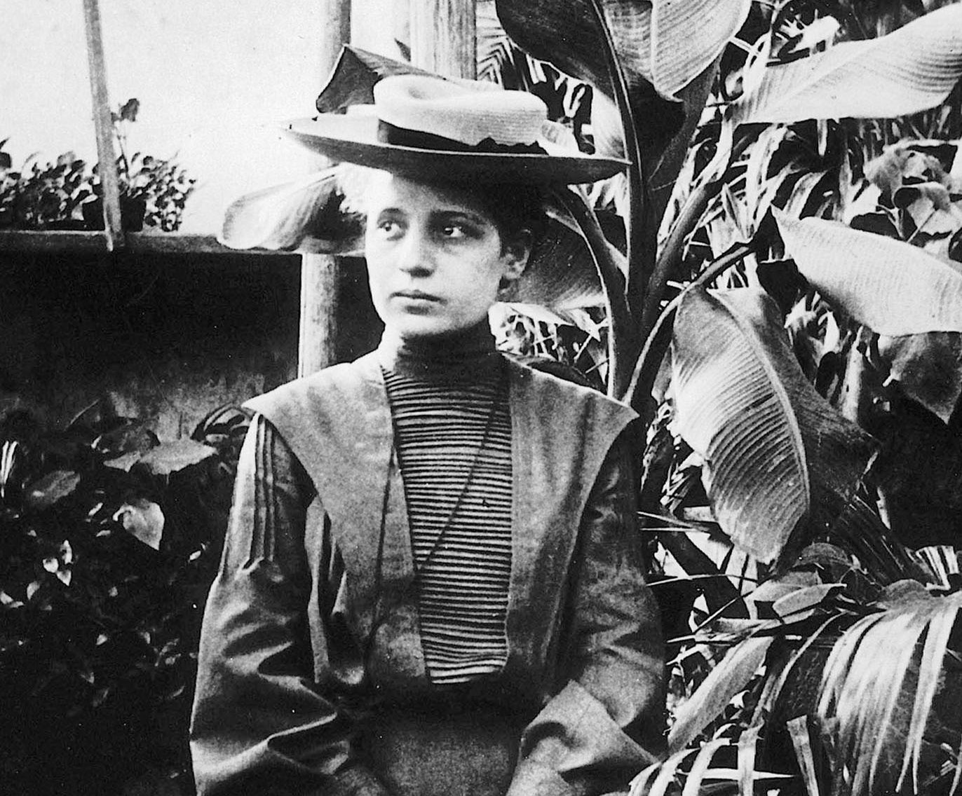 Ein Schwarzweißfoto zeigt eine Frau in historischer Kleidung und mit Hut, die in einem Garten mit großen Blättern neben einem Fenster steht.