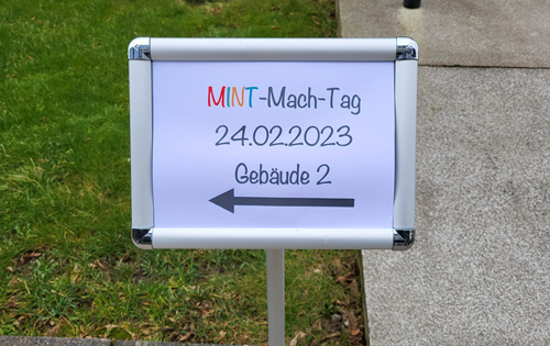 Das Bild zeigt ein Schild, auf dem steht "MINT-Mach-Tag 24.02.2023 Gebäude 2". Ein Pfeil zeigt den Weg zum Gebäude.