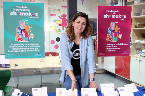 Das Foto zeigt Nuria Robles. Sie steht zwischen zwei Postern, auf denen einmal auf Englisch und einmal auf Spanisch steht: Dieses Labor ist Teil des shemakes-Netzwerks. Wir stärken die weiblichen Innovatorinnen von morgen in der nachhaltigen Modeindustrie durch Inspiration, Fähigkeiten und Netzwerke.