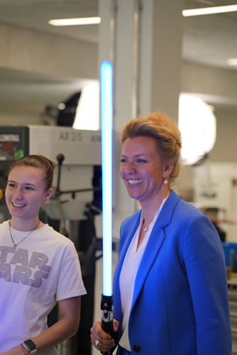 Ministerin Ina Brandes lächelt breit. Sie hat ein blau leuchtendes Lichtschwert in der Hand. Neben ihr steht Schülerin Amelie, die ebenfalls lächelt.
