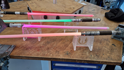 Das Foto zeigt vier im Kurs hergestellte Lichtschwerter, die auf aus Acrylglas im Kurs gefertigten Ständern liegen. Die Klingen leuchten in den Farben orange, grün, pink und orange.