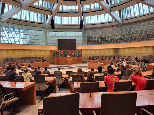 Das Foto zeigt den Plenarsaal im Landtag von NRW. Im Saal verteilt sitzen junge und ältere Menschen. Am Rednerpult steht eine Frau.