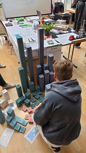 Das Foto zeigt verschiedenfarbige Bauklötzchen, die vor einem Messestand aufgebaut sind. Die Klötzchen repräsentieren verschiedene Bausteine, die CO2-Ausstoß verursachen. Aus ihnen wird ein persönlicher Klimaturm aufgebaut.