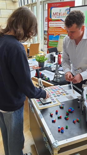 Das Foto zeigt zwei Menschen am Stand von MExLab ExperiMINTe, die mit einer Stanzmaschine bunte Buttons aus Papier ausstanzen.