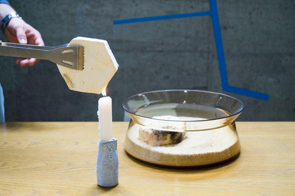 Das Foto zeigt ein Stück Pilzmaterial, das über die Flamme einer Kerze gehalten wird. Dahinter steht eine Glasschale mit Sand, in der sich ein weiteres, bereits angekokeltes Stück Pilzmaterial befindet.