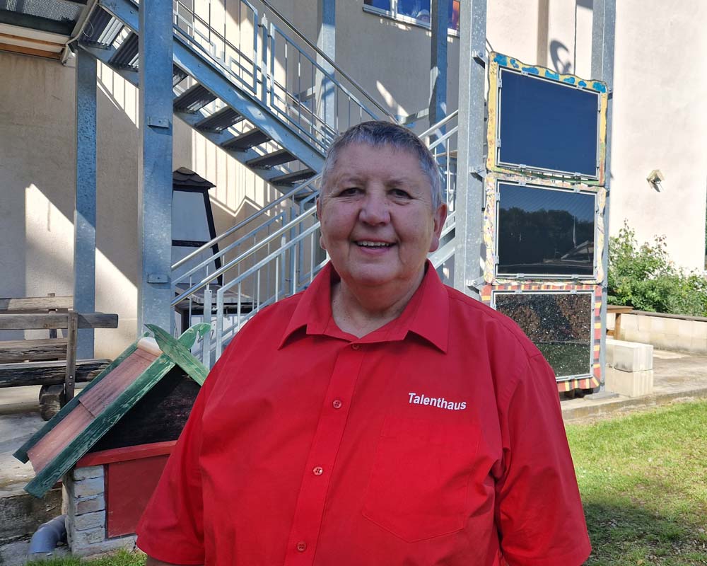 Das Foto zeigt Karin Ressel. Sie trägt ein rotes Hemd auf dem Talenthaus steht und lächelt in die Kamera.