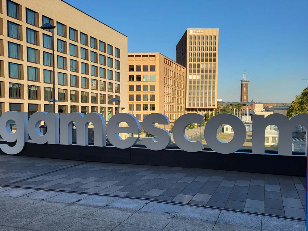 Das Foto zeigt den Schriftzug "gamescom" in großen, grauen Buchstaben vor der Skyline der Kölnmesse und einem strahlendblauen Himmel.