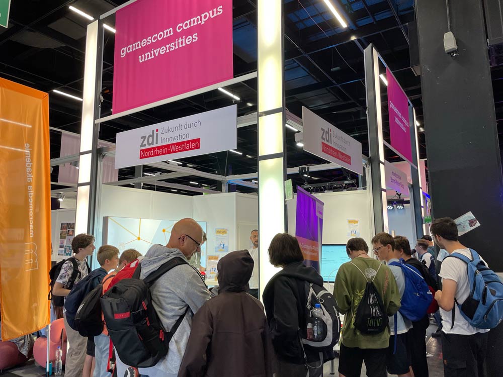 Das Foto zeigt den Stand von zdi.NRW auf der Gamescom. Auf einem Schild über dem Stand steht "gamescom campus universities". Um den Stand steht eine Menge aus Menschen unterschiedlichen Alters.