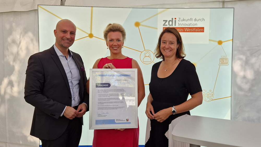 Das Foto zeigt Landrat Ali Doğan, Ministerin Ina Brandes und Regierungspräsidentin Anna Katharina Bölling. Die Ministerin hält eine Urkunde zur Ernennung des zdi-Schüler:innenlabors in der Hand. Die drei stehen vor einer Leuchtwand, auf der zdi - Zukunft durch Innovation Nordrhein-Westfalen steht.