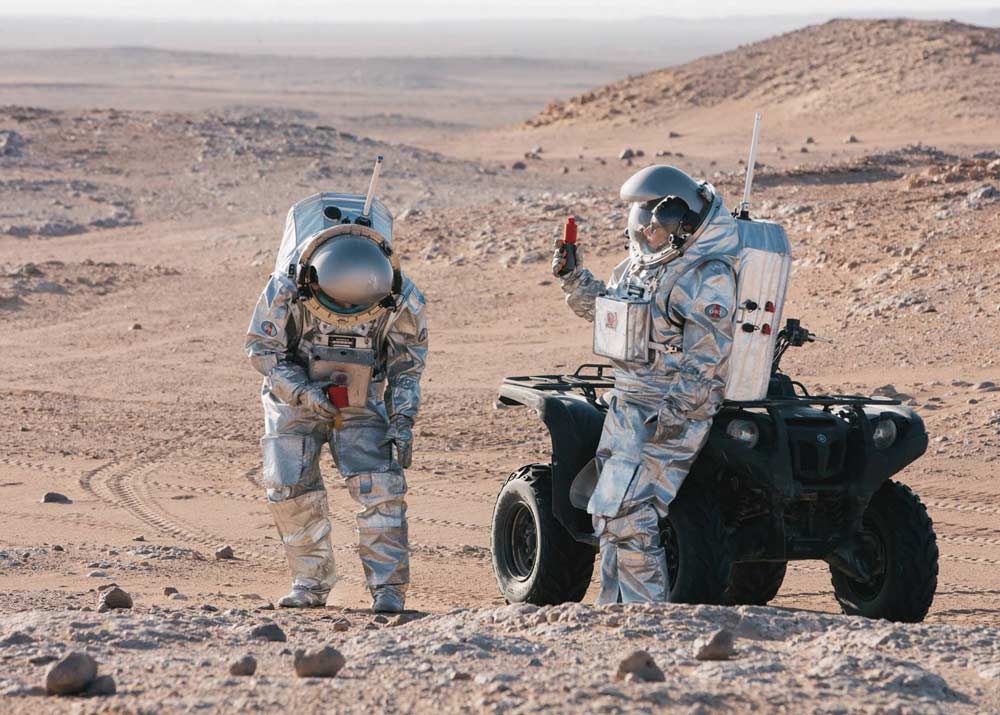 Zwei Astronauten stehen neben einem Geländefahrzeug in der Wüste.