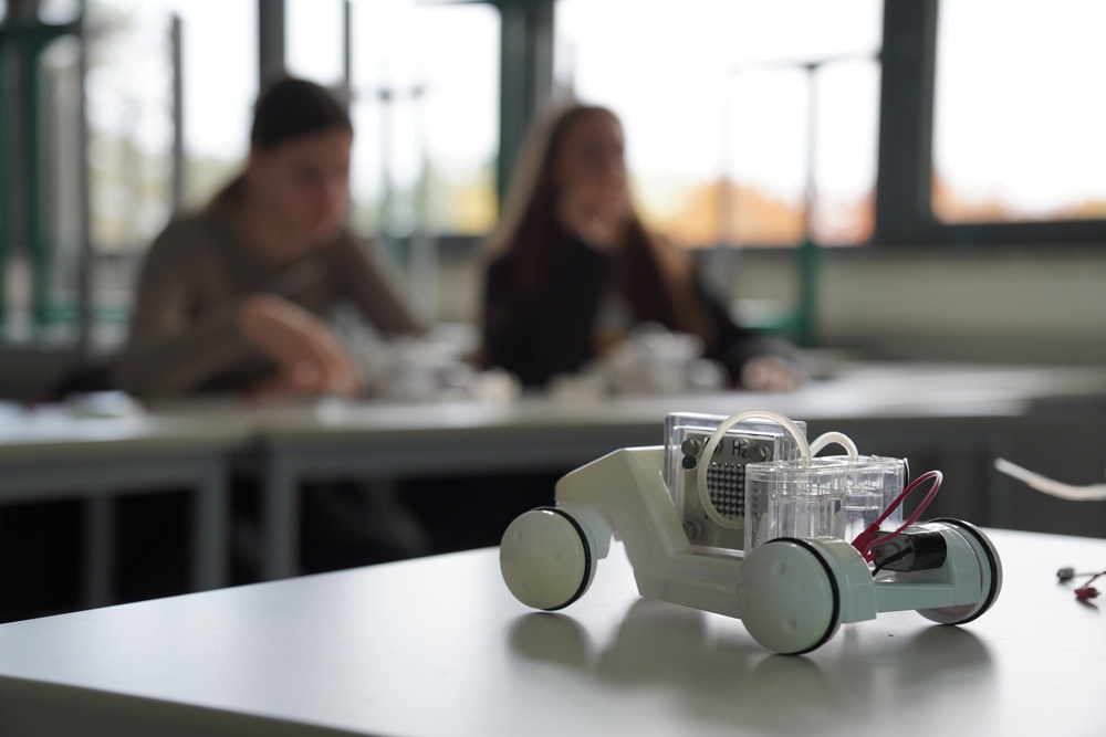 Ein Spielzeugauto, das beim zdi-Mädchen-Camp auf einem Tisch in einem Klassenzimmer steht. Im Hintergrund sitzen zwei Mädchen.