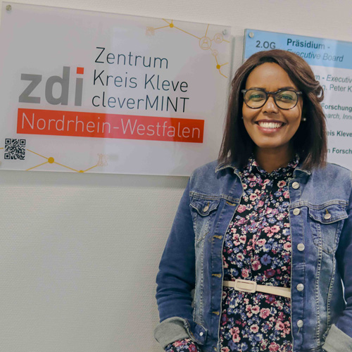 Phyllis Ndugire steht lächelnd vor einem Schild, auf dem steht: zdi-Zentrum Kleve cleverMINT Nordrhein-Westfalen.
