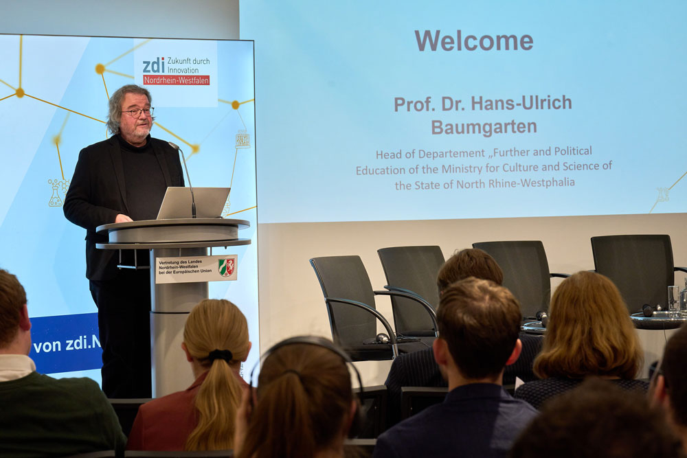 Eine Gruppe von Menschen sitzt in einem Raum und hört den Grußworten von Prof. Dr. Hans-Ulrich Baumgarten zu, der über zdi und MINT spricht.