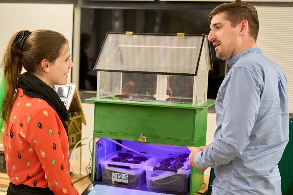 Eine Gruppe von Menschen steht um einen Tisch und schaut auf eine Indoor-Farm. Hier präsentiert das zdi-Schüler:innenlabor Hamm die Ergebnisse der zdi-Science League zur MINT-Förderung.