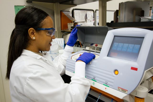 Eine Frau bzw. Forscherin schaut durch ein Mikroskop. Neben dem Mikroskop steht ein weiteres kleineres wissenschaftliches Instrument.