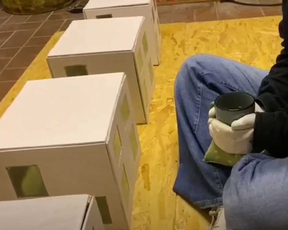 Vier Hausmodelle stehen in einer Reihe. Davor sitzt ein Mensch im Schneidersitz mit einer Tasse in der Hand.