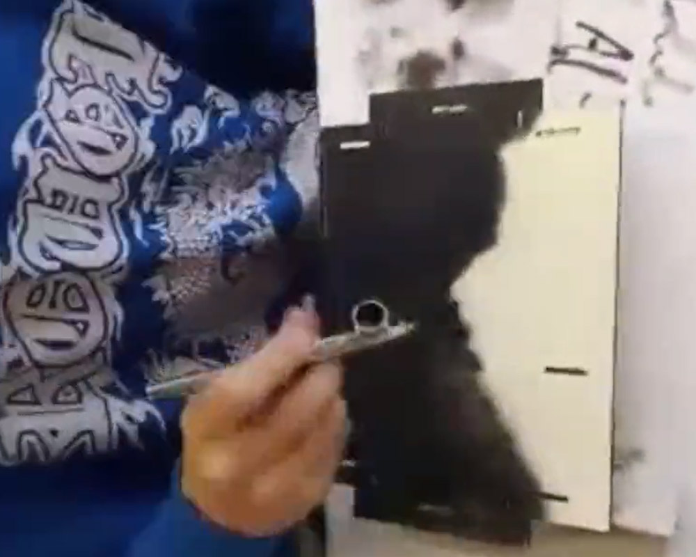 Ein Mensch in einem blauen Pullover besprüht ein Stück Holzplatte mit schwarzer Airbrush-Farbe.