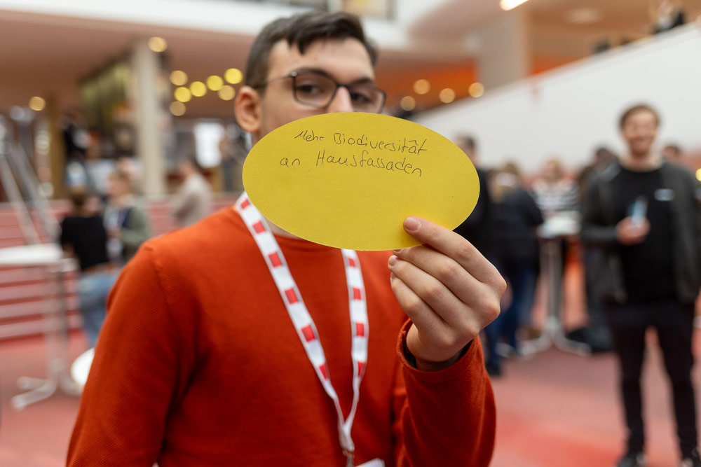 Ein Mann hält ein gelbes Papier mit einer Notiz darauf hoch. Die Notiz besagt: Mehr Biodiversität an Hausfassaden.