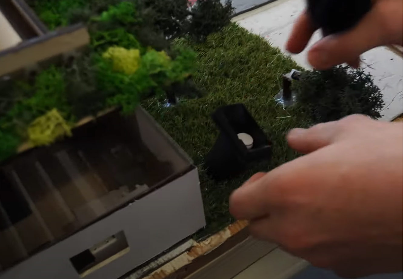 Eine Hand justiert eine Miniaturszene mit Bäumen und einer Mülltonne auf dem Boden.