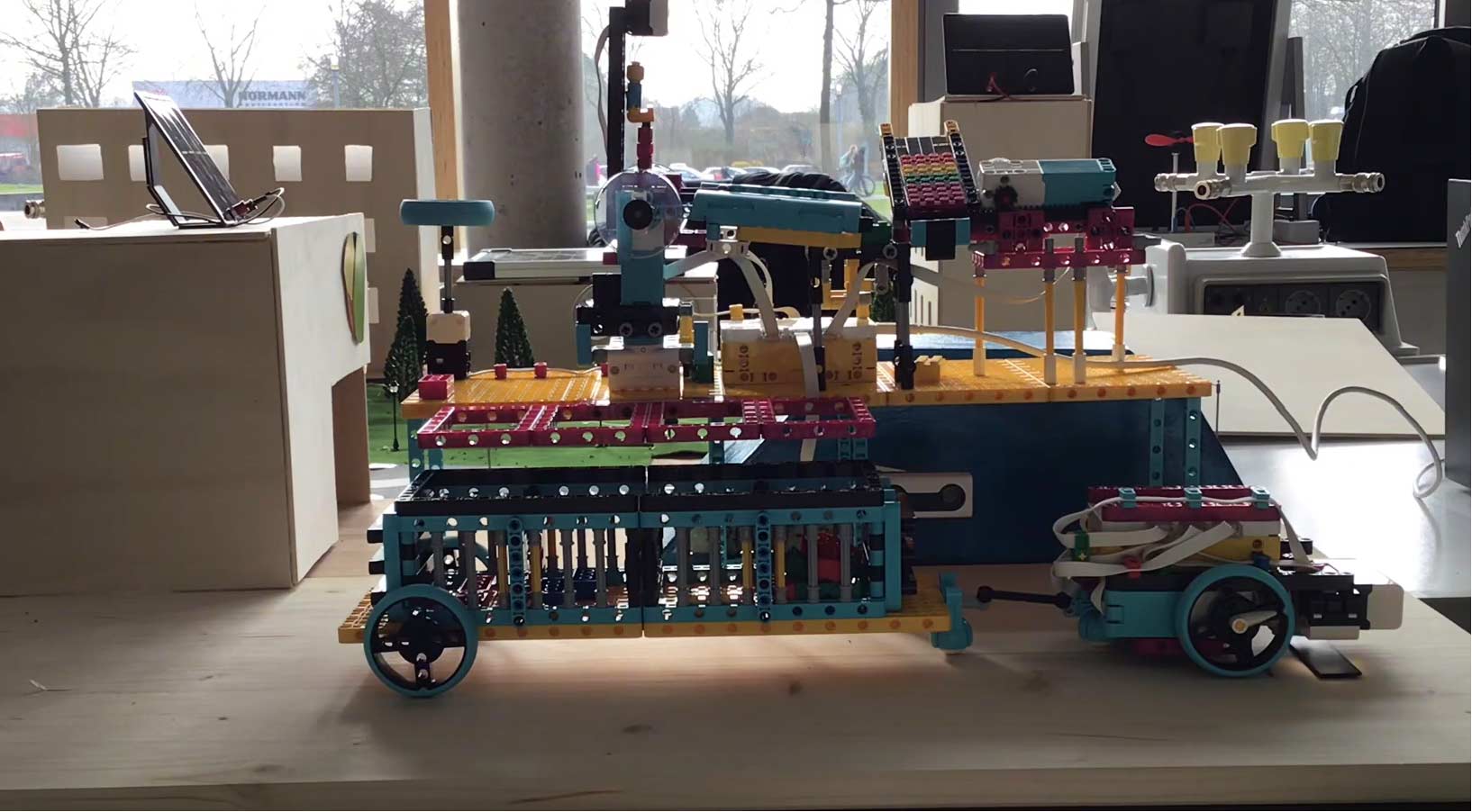 Eine mehrfarbige Robotermaschine, die auf einem Holztisch ausgestellt ist, im Hintergrund sind Modellhäuser zu sehen.