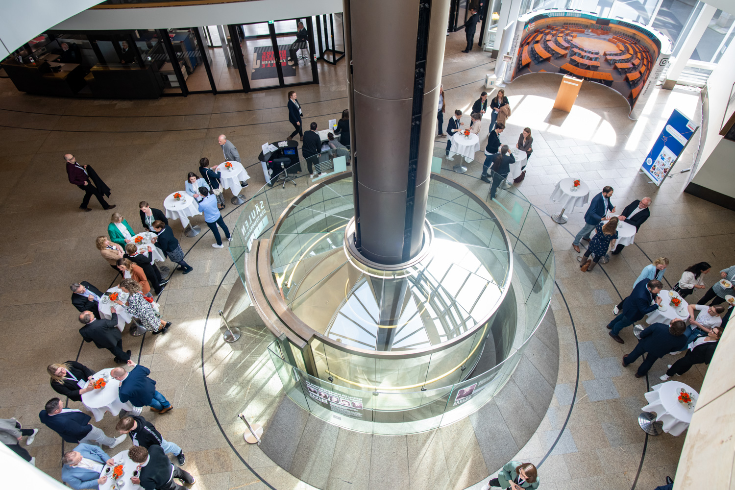Das Bild zeigt eine Luftaufnahme eines belebten Innenatriums. Man sieht Menschen, die an Tischen sitzen und eine markante zentrale Glaszylinderstruktur ist sichtbar. Das Atrium wurde mit dem zdi-Qualitätssiegel ausgezeichnet.