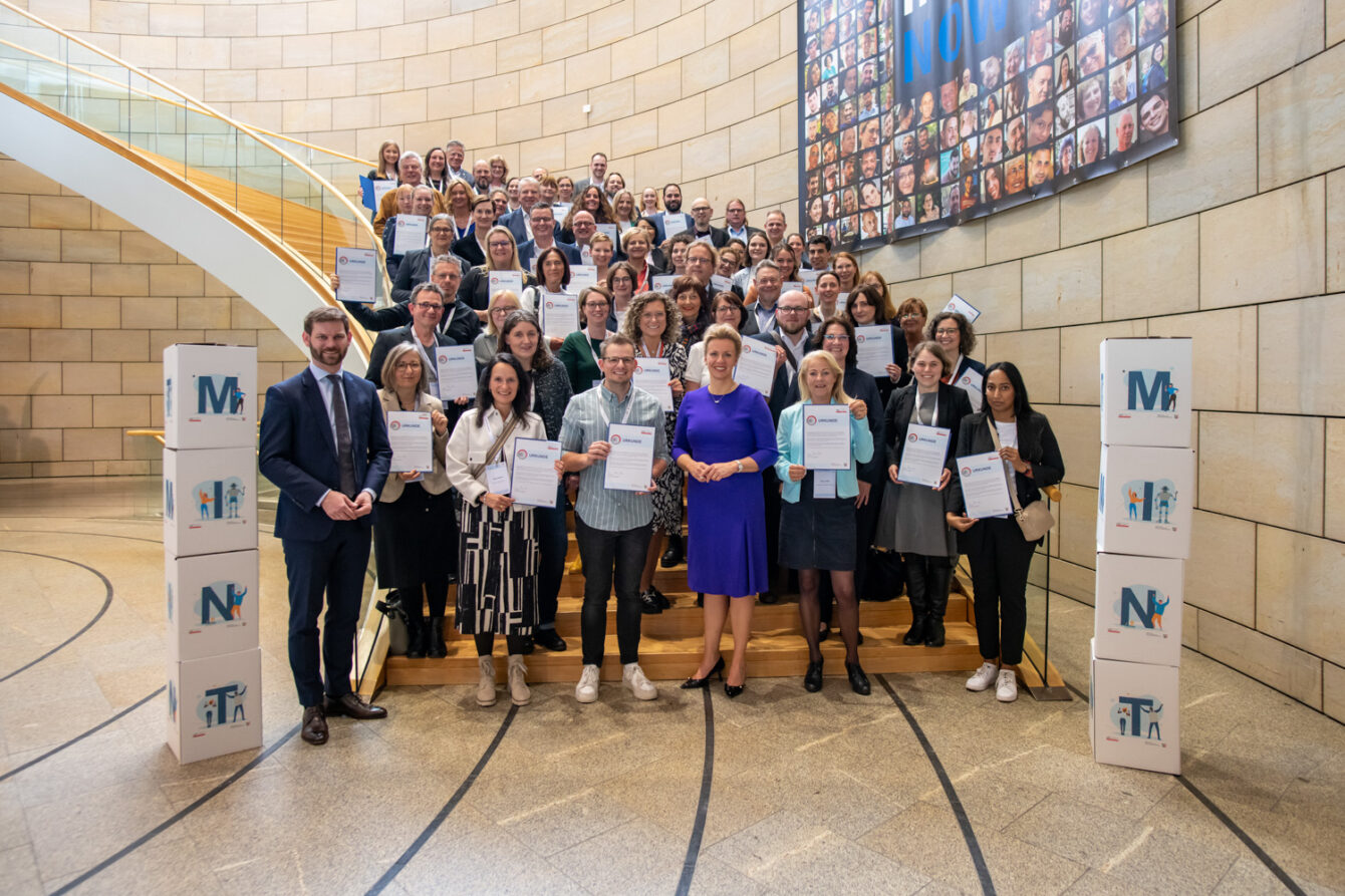 In einem modernen Atrium präsentiert eine Gruppe von Fachleuten zdi-Qualitätssiegel-Zertifikate. Sie stehen versetzt zueinander, im Hintergrund ist eine Treppe zu erkennen.