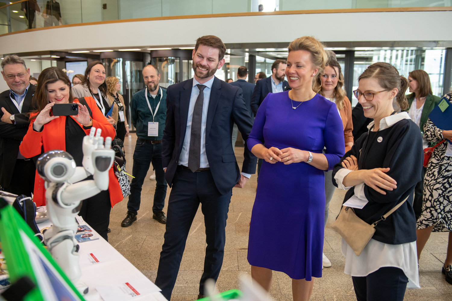 Auf einer zdi-Qualitätssiegel-Konferenz sieht man eine Gruppe von Fachleuten lächelnd mit einem kleinen weißen Roboter interagieren. Zu der Gruppe gehört auch eine blau gekleidete Frau.