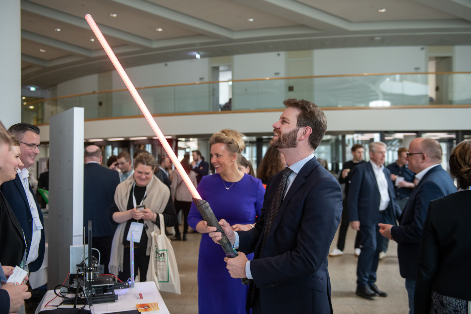 Ein Mann im Anzug demonstriert aufgeregt ein rosa Lichtschwert mit zdi-Qualitätssiegel. Eine Frau im blauen Kleid beobachtet lächelnd eine Indoor-Veranstaltung. Schaulustige sind anwesend.