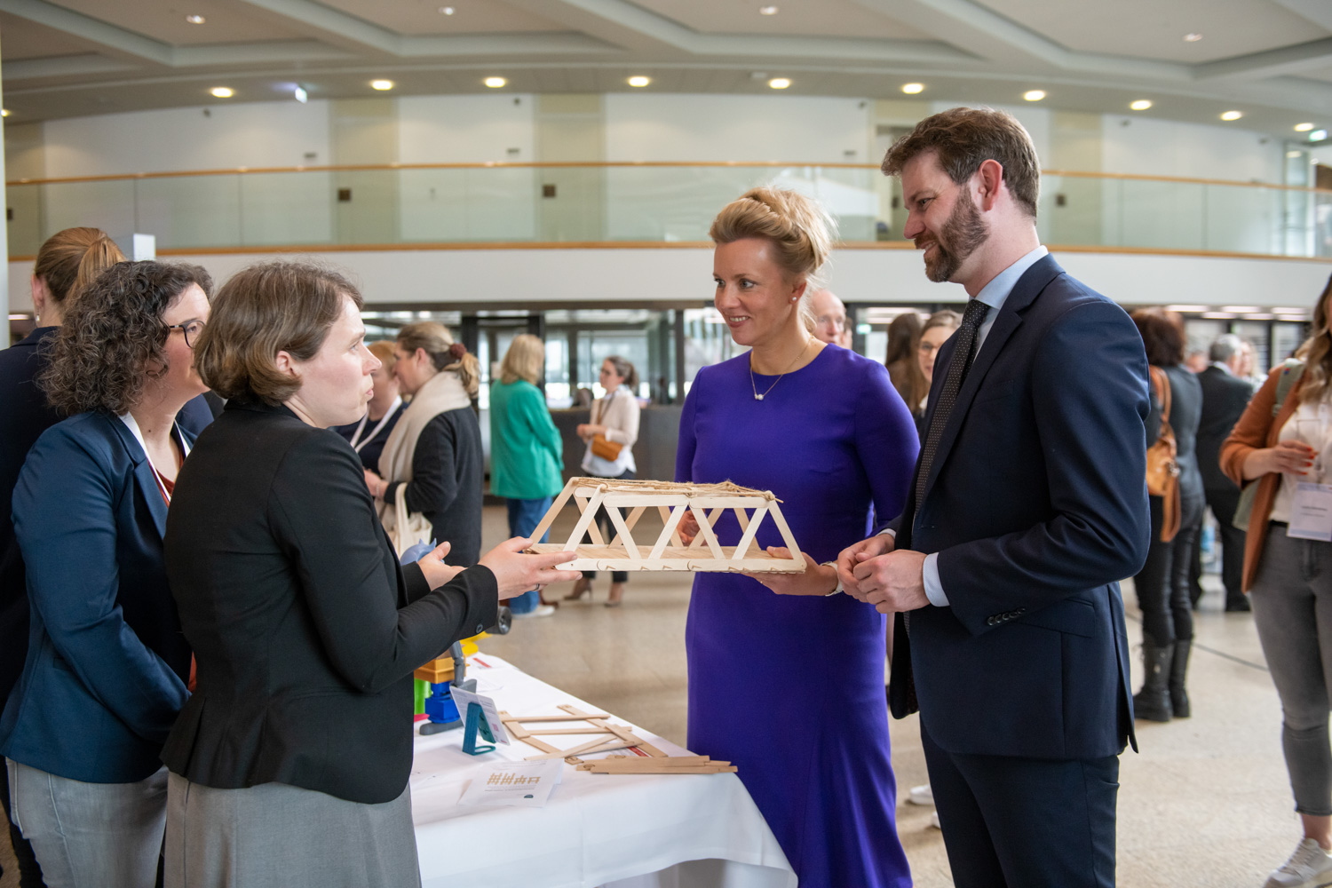 Auf einer Konferenz betrachten zwei Teilnehmer, ein Mann und eine Frau in formeller Kleidung, ein Holzbrückenmodell mit zdi-Qualitätssiegel. Sie unterhalten sich mit anderen Teilnehmern.