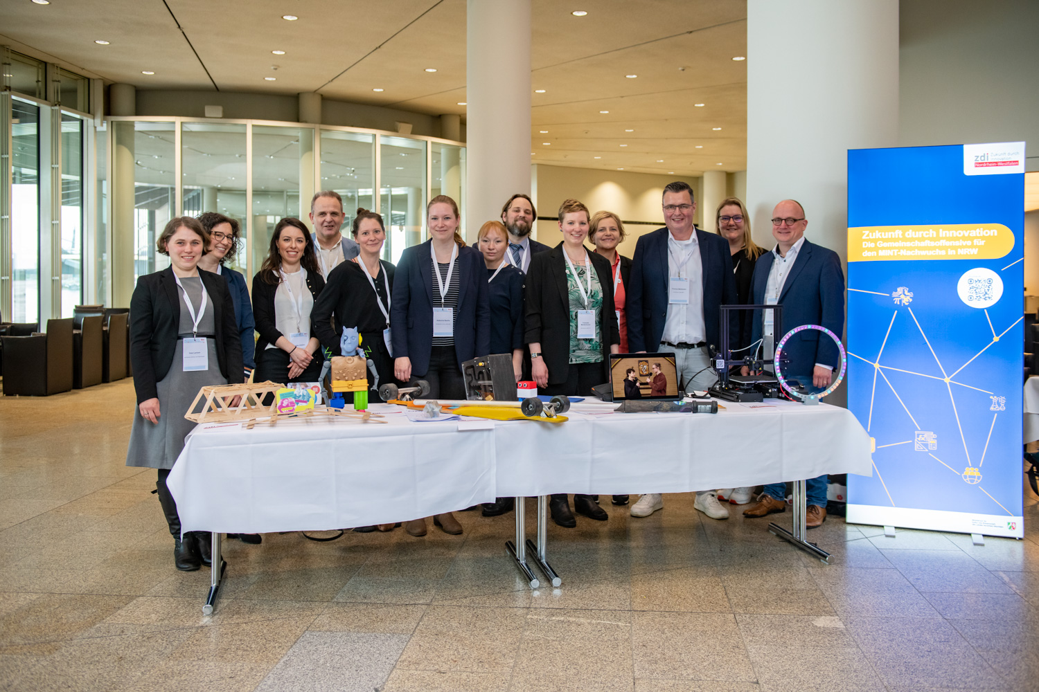Eine lächelnde Gruppe von Fachleuten steht an einem Konferenzstand. Sie sind umgeben von verschiedenen technologischen und lehrreichen Exponaten wie Modellen und elektronischen Geräten. Diese tragen alle das zdi-Qualitätssiegel.