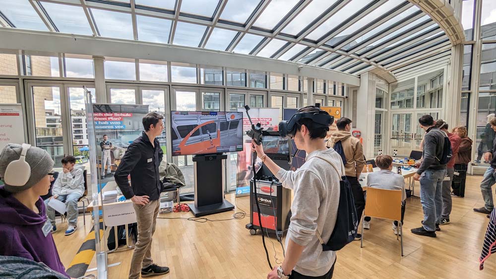 In einem Gebäude mit Glasdach nehmen Einzelpersonen an der Technologiepräsentation des Zukunftstags 2024 teil. Sie nehmen an verschiedenen Technologiepräsentationen teil und nutzen Virtual-Reality-Geräte.