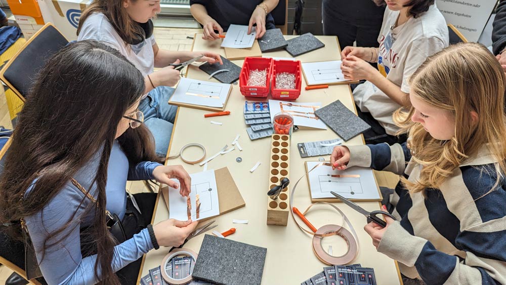 Auf dem Bild sieht man Schülerinnen und Schüler an einem Werkstatttisch für den Zukunftstag 2024 bei einem Elektronikprojekt. Sie bauen Schaltkreise zusammen und diskutieren ihre Arbeit.