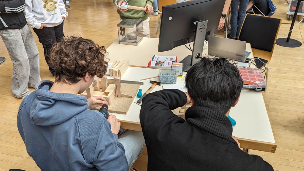 Zwei Personen sitzen an einem Werkstatttisch und bauen während des Zukunftstags 2024 Holzmodelle zusammen. Sie sind umgeben von Laptops und Werkzeug. Im Raum befinden sich auch mehrere Zuschauer.