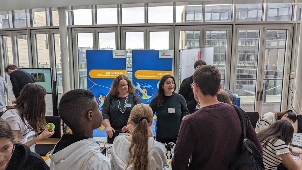 Eine Gruppe von Personen unterhält sich an einem belebten Stand der Zukunftstag-Konferenz 2024. Im Hintergrund sind Werbebanner zu sehen.