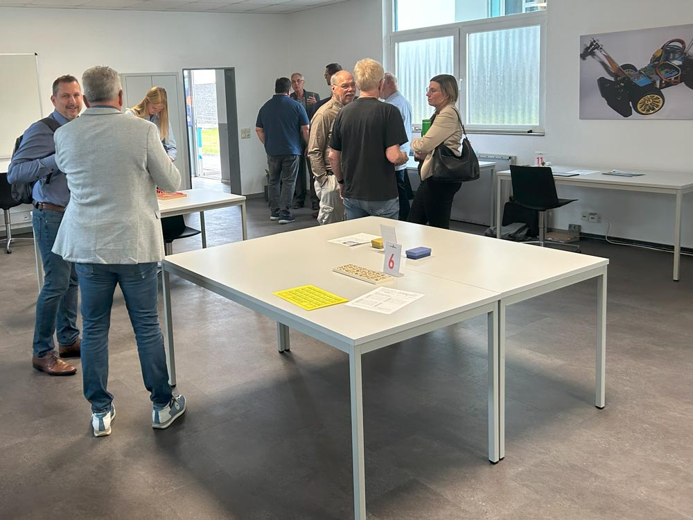 In einem hell erleuchteten Raum mit großen Fenstern im PhänomexX Datteln steht eine Gruppe von Menschen an Tischen und unterhält sich. Auf einigen Tischen liegen Papiere und Broschüren. An der Wand hängt ein Spielzeugauto.