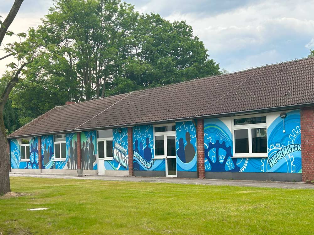 Das Bild zeigt ein einstöckiges Gebäude mit einem Wandgemälde aus blauen Zahnrädern, Personensilhouetten und verschiedenen Grafiken an der Außenseite. „PhänomexX Datteln“ ist von Bäumen und einer Rasenfläche umgeben.