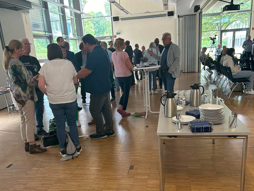 Eine Gruppe von Menschen steht und unterhält sich in einem Innensaal, während andere sitzen. In der Nähe steht eine Kaffeestation mit Tassen, Tellern und Thermoskannen. Große Fenster lassen Tageslicht herein. Die Veranstaltung mit dem Namen PhänomexX Datteln versammelt neugierige Köpfe in einer warmen und einladenden Umgebung.