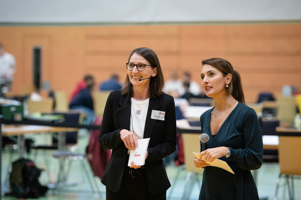 Zwei Frauen stehen in einer Sporthalle. Die rechte Frau ist Staatssekretärin Gonca Türkeli-Dehnert. Sie hat ein Mikrofon in der Hand.