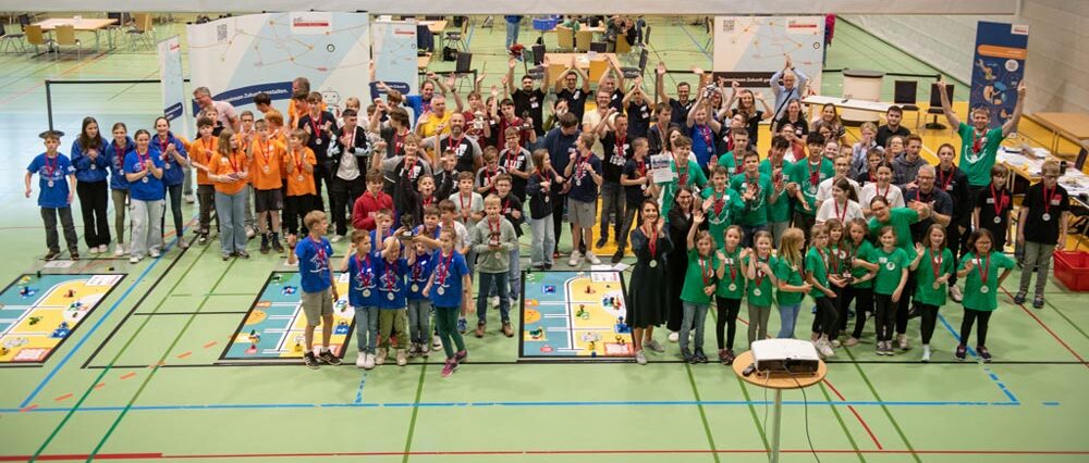 Eine große Gruppe von Menschen, darunter Kinder und Erwachsene in bunten Hemden, versammelt sich in einer Turnhalle zum zdi-Roboterwettbewerb. Die Teilnehmer halten stolz Medaillen und Trophäen hoch. Am Boden sind mehrere Robotermatten zu sehen.