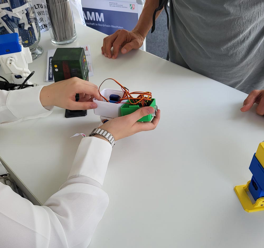 Beim NRW-Tag 2024 führt eine Person im weißen Hemd einer anderen Person an einem Tischdisplay ein kleines grün-weißes elektronisches Gerät mit Kabeln vor.