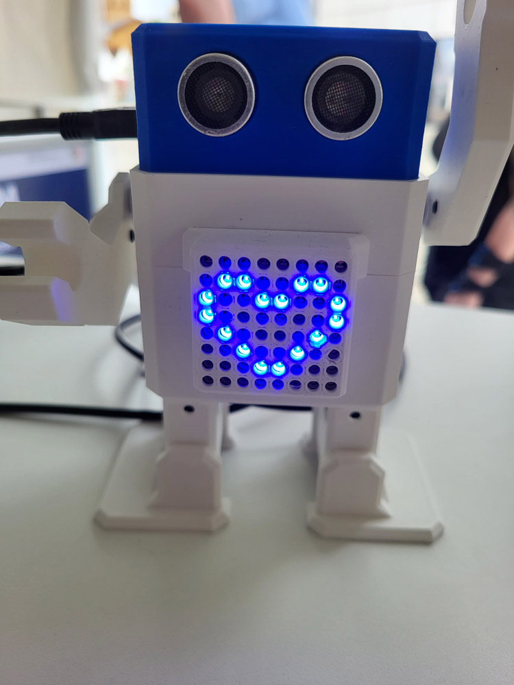 Ein kleiner weiß-blauer Roboter winkt und zeigt auf seiner Vorderseite ein Herz aus blauen LED-Lichtern.