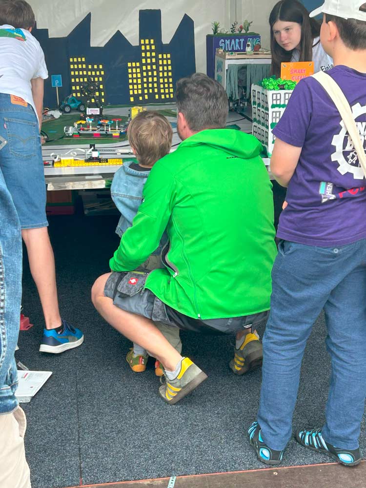 Ein Mann in einer grünen Jacke kniet neben einem kleinen Jungen an einem Tisch mit einem Stadtmodell. Andere Leute stehen herum und beobachten die komplizierte Kreation.
