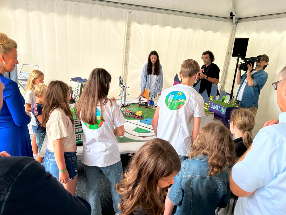 Beim NRW-Tag 2024 versammeln sich Kinder und Erwachsene um einen Tisch mit einem Miniatur-Stadtmodell in einem Zelt. Einige schauen zu, während andere mit dem Exponat interagieren. Eine Person filmt die Szene.